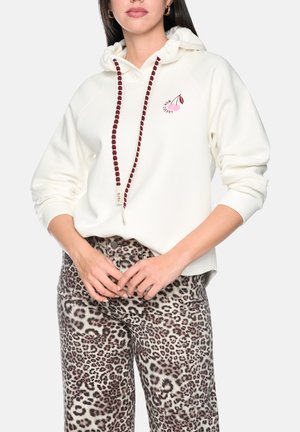 Frau trägt weißen Kapuzenpullover mit Kirschgrafik und bordeauxfarbenen Schnürsenkeln, kombiniert mit Leopardenmusterhose, steht mit verschränkten Händen.