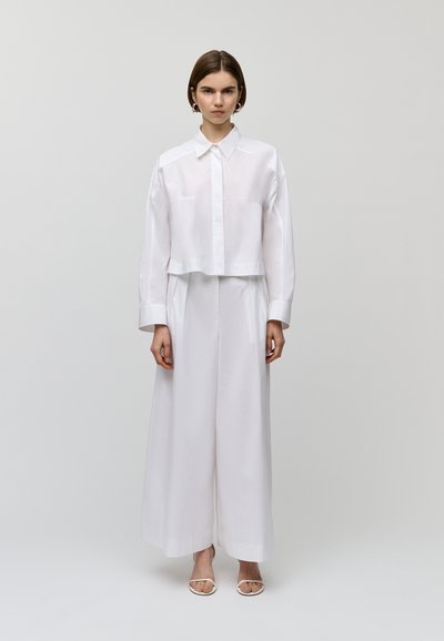 Camicia bianca cropped con colletto, abbinata a pantaloni bianchi a gamba larga e sandali bianchi con cinturini, su uno sfondo neutro.