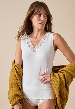 Donna che indossa un top senza maniche bianco con rifiniture in pizzo e biancheria intima coordinata, con uno scialle giallo senape dalla texture drappeggiato sulle braccia.