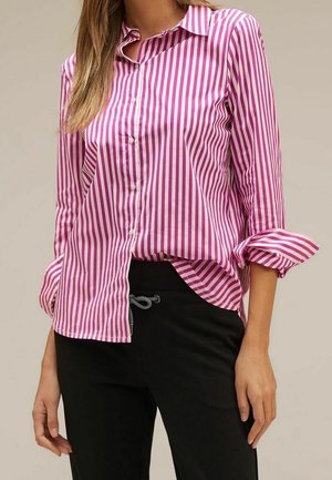 Frau trägt ein rosa-weiß vertikal gestreiftes Hemd mit hochgekrempelten Ärmeln und schwarze Hosen mit Kordelzug in der Taille.