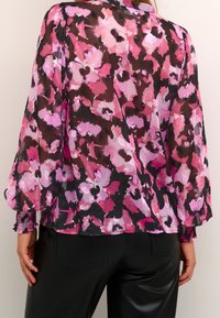 Kvinde iført en gennemsigtig bluse med pink, lilla og sort blomstermønster og sorte tætsiddende bukser, vist fra ryggen.