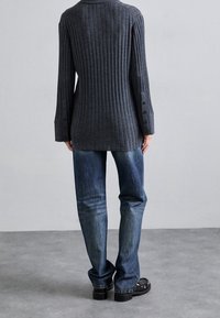 Person trägt ein dunkel geripptes Langarmshirt mit geknöpften Manschetten, blaue Jeans und schwarze Schuhe und steht auf einem grauen Boden vor einer einfarbigen Wand.