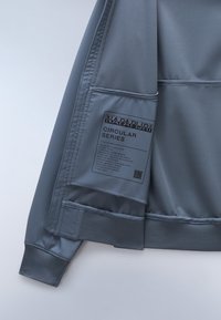 Hellgraue Jacke aus strukturiertem Material, mit gerippten Bündchen und einem bedruckten Etikett, das die "Circular Series" beschreibt. Glattes, sportliches Design.