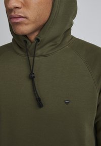 Hombre con sudadera verde oliva con cordones negros y un pequeño logo triangular en el pecho.