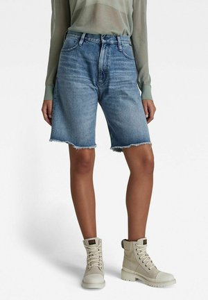 Personne portant un short bermuda en denim bleu clair avec des ourlets effilochés et des bottines beiges à lacets, sur un fond uni.