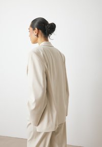 Beige tailored blazer with dropped shoulders; smooth fabric, minimal stitching details, and a sophisticated cut. Pair of matching trousers visible.