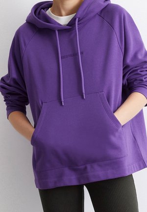 Personne portant un sweat à capuche violet avec une poche avant et des cordons, mains placées dans la poche, visible du cou aux hanches.