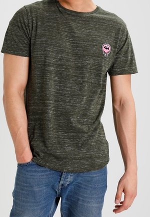 Mann trägt ein dunkelgrünes, meliertes Kurzarm-T-Shirt mit einer kleinen pinkfarbenen Grafik schmelzender Lippen auf der linken Brust, kombiniert mit blauen Jeans.