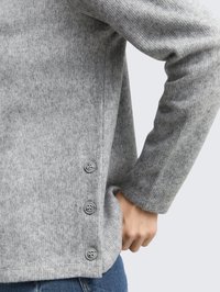 Maglione grigio in maglia con una texture morbida, dotato di una tasca laterale e quattro bottoni grigi lungo l'orlo per una vestibilità regolabile.