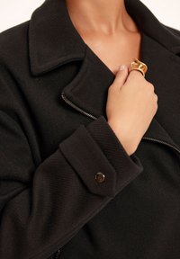 Czarna kurtka wykonana z teksturowanej tkaniny, z dużym kołnierzem, srebrnym zamkiem błyskawicznym i zapinanymi mankietami. W tym również złoty pierścień noszony na ręce.