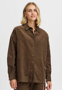 Camisa de pana marrón con cierre de botones en la parte delantera, mangas largas y un corte relajado. Presenta un cuello clásico y una textura sutil.