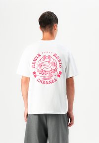 Hvit bomulls t-skjorte med et rødt grafisk design av en frosk med volangskjørt, omgitt av tekst. Korte ermer, avslappet passform.