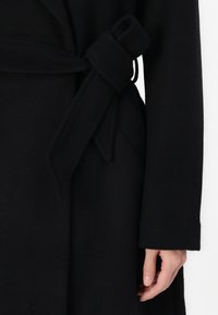 Manteau en laine noir avec une ceinture nouée à la taille, à manches longues et à la finition douce et texturée. Le tissu présente un aspect lisse.