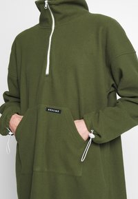 Grön fleece-tröja med hög krage, halv dragkedja framtill, kenguruficka, elastiska muddarna och en etikett med texten "RÉSUMÉ."