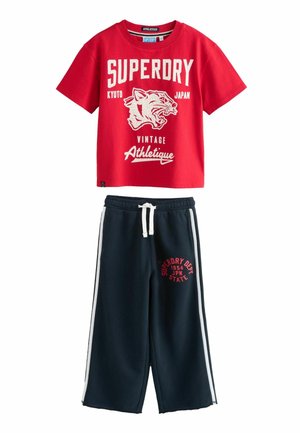 Röd bomullst-shirt med kort ärm och vitt grafiskt tryck samt rundad halsringning, tillsammans med marinblå träningsbyxor med vita sidostripes och logotyp.