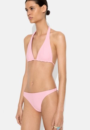 Kobieta w jasnoróżowej górze bikini z wiązaniem na szyi i pasujących do niej dołach bikini, stojąca na jednolitym białym tle.