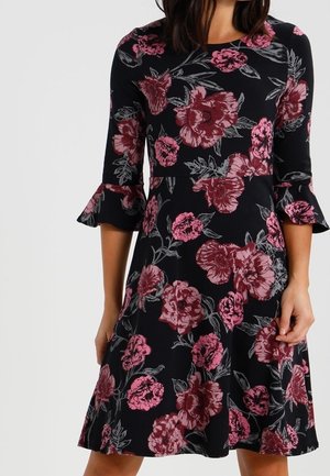 Czarna sukienka do kolan z dużym różowym kwiatowym nadrukiem, rękawy o długości do łokcia i falbaniaste mankiety, noszona przez osobę.