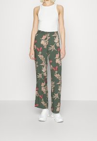 Zielone kwiatowe spodnie z różowymi i czerwonymi motylkami, uszyte z lekkiego materiału; w zestawie z białym topem na ramiączkach i białymi trampkami.