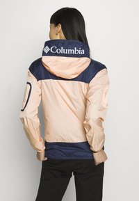 Lättviktsjacka med huva i persika och marinblå, med elastiska ärmslut, en figursydd nederkant och en logotyp på baksidan. Slät, vattenavvisande tyg.