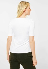 Bílý krátký rukáv tričko s kulatým výstřihem, slim fit, a hladkým bavlněným povrchem, nošené s tmavě zelenými kalhotami.
