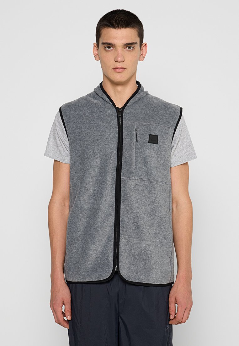Gilet sans manches gris avec une fermeture éclair à l'avant, des finitions noires contrastantes et une petite poche poitrine. Superposé sur une chemise à manches courtes grise.