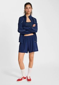 Giacca sportiva blu navy con bordi bianchi, abbinata a canottiera e pantaloncini blu navy coordinati. La modella indossa sneaker rosse e calzini bianchi alla caviglia.