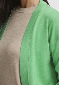 Kobieta nosząca beżowy sweter pod jasnoniebieskim kardiganem, pokazana od klatki piersiowej do szyi, z długimi ciemnymi włosami widocznymi.