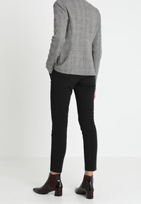 Una persona è in piedi con la schiena rivolta verso la macchina fotografica, indossando un blazer grigio a righe, pantaloni neri e stivaletti lucidi.