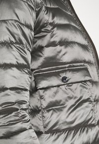 Grå dunjakke med en glat tekstur, der har vandret quilting, en klaplomme sikret med en sort knap og en lynlåslukning foran.