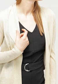 Beige kofta med en jämn textur, öppen framsida och lång ärmar. Bärs över en svart jumpsuit med ett brett bälte och V-ringning.