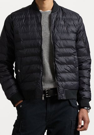 Mann trägt eine schwarze gesteppte Bomberjacke über einem grauen Pullover, schwarze Hosen und einen schwarzen Gürtel, Hände in den Taschen und eine Armbanduhr sichtbar.