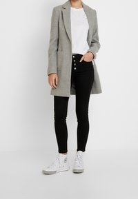 Šedý dlouhý kabát, bílý tričko, černé slim jeans s knoflíkovými detaily a bílé tenisky s vysokým střihem a černobílou kostkovanou podrážkou.