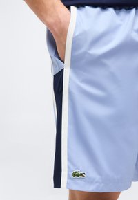 Ανοιχτό μπλε αθλητικό σορτς με λευκές και ναυτικές λωρίδες στις πλευρές και πράσινο λογότυπο κροκόδειλου κοντά στο τελείωμα, φορεμένο με το χέρι στην τσέπη.