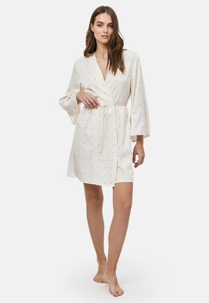 Vrouw draagt een crèmekleurige wikkeljurk met kleine zwarte stippen en brede mouwen, staat op blote voeten tegen een effen achtergrond.