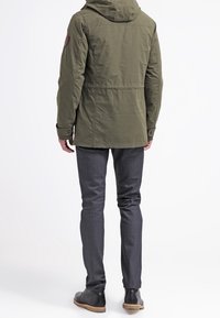 Olive green parka with a hood, featuring a brown leather patch on the shoulder, paired with dark gray slim-fit jeans and black boots.