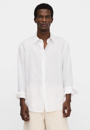 Man draagt een wit overhemd met lange mouwen, opgeknipte manchetten en lichte beige broek, staand tegen een effen achtergrond.