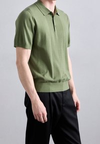 Hombre vestido con una camiseta polo de manga corta verde, ligeramente metida en unos pantalones negros, de pie contra un fondo claro y liso gris.