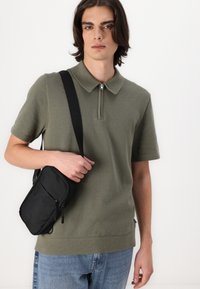 Olivgrünes Poloshirt mit Reißverschlusskragen, strukturiertem Stoff, kurzen Ärmeln, kombiniert mit einer kleinen schwarzen Umhängetasche und blauen Jeans.