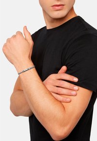Pulsera trenzada de plata con una placa metálica plana, llevada en una muñeca, exhibida sobre una camiseta negra. Textura suave y diseño sencillo.