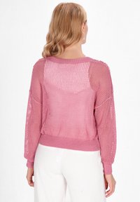 Strickpullover in Hellrosa mit einem Netzdesign, langen Ärmeln, gerippten Manschetten und Rückendetails, kombiniert mit weißen Hosen.