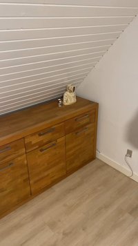 Houten dressoir met metalen handgrepen onder een schuin wit geveeld plafond; twee kleine beeldjes erop; witte muur en een stopcontact in de buurt.