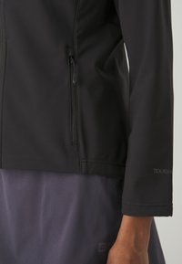 Giacca soft-shell nera con tasca con zip sulla vita sinistra, caratterizzata da una texture liscia e un logo del marchio visibile sulla manica.