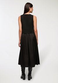 Czarny top bez rękawów o skróconym fasonie, zestawiony z długą, plisowaną czarną spódnicą. Modelka ma na sobie czarne kozakami na przysłoniętych kolanach.