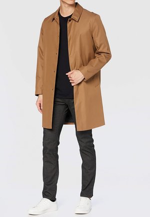 Manteau long beige avec un col, des boutons sur le devant et des poches latérales, assorti à un pantalon sombre et des baskets blanches. Texture lisse et design moderne.