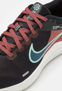 Svart idrottssko med röda snören, texturerad tygöverdel, reflekterande aqua Nike-swoosh och en vit mellansula. Anmärkningsvärd branding på tungan.