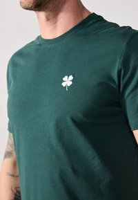 Donkergroen katoenen t-shirt met een klein wit vierbladig klavertje geborduurd op de linkerborst. Eenvoudig crew neck ontwerp.