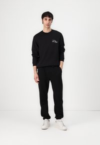 Czarna bawełniana bluza z logo z przodu oraz dopasowane czarne dresy zwężane. Model w białych sneakersach, stojący na tle jednolitym.
