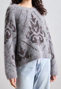 Žena oblečená v huňatém šedém svetru s tmavými abstraktními vzory a světle modrými džínami s vysokým pasem, stojící na jednoduchém pozadí.