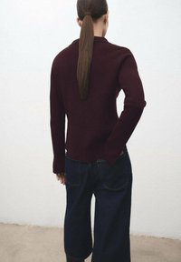 Burgundska pletena pulover s dolgimi rokavi, rebrastimi manšetami in zaobljenim robom, v kombinaciji s širokimi temnimi denim culotte hlačami. Prikazan zadnji pogled.