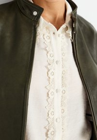 Chaqueta de cuero verde oscuro con cremallera frontal, que cuenta con un alto cuello. Debajo, una blusa blanca con un borde de encaje texturizado y botones.